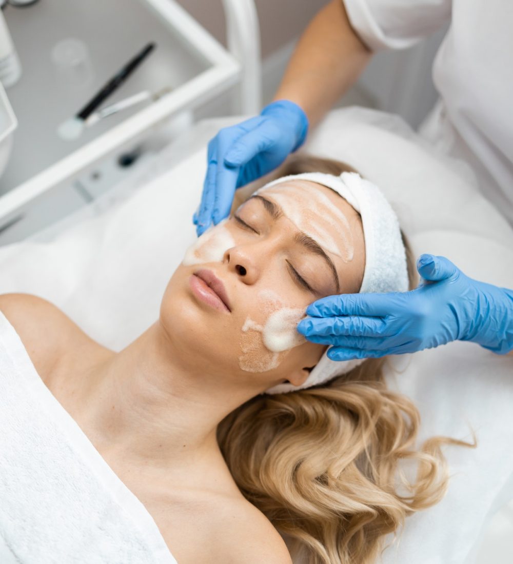 Beautician doctor in blue gloves is applying facial cleansing foam or cream on woman's face