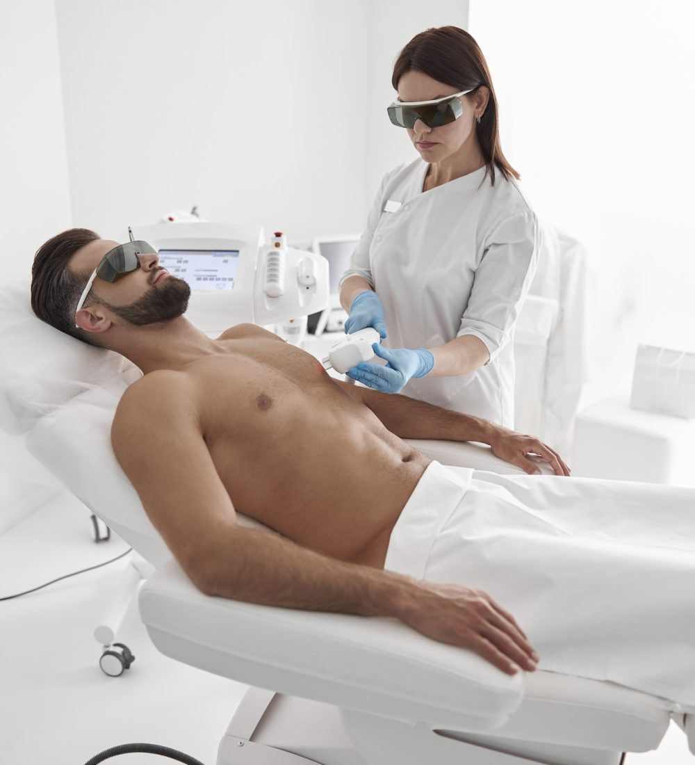 Young master with goggles conducts laser epilation to bearded man client lying on couch in clinic